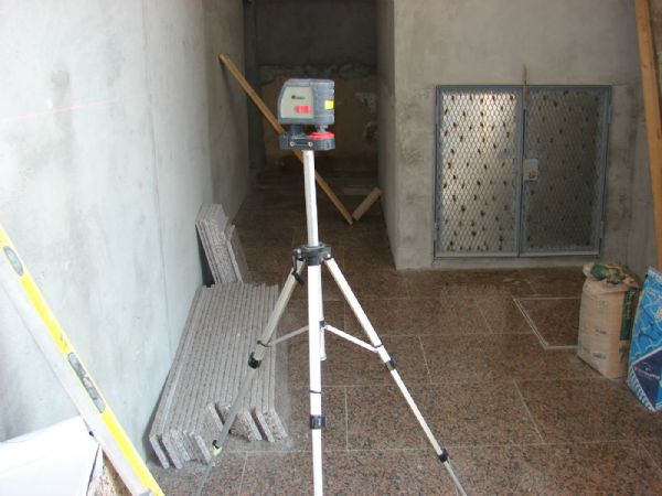 En la imagen se observa el granito colocado en el suelo, la puerta de armario de contadores de agua, realizada a medida, con acero, dejando hueco para rellenar con la piedra y soldando chapa deploy para el agarre de las masas. Se puede observar el laser con el que se estaban marcando los niveles para colocar el friso de granito